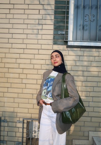 Joven mujer con hijab negro lleva una chaqueta gris, camiseta gráfica, pantalones blancos y lleva un gran bolso verde al hombro contra una pared de ladrillos.