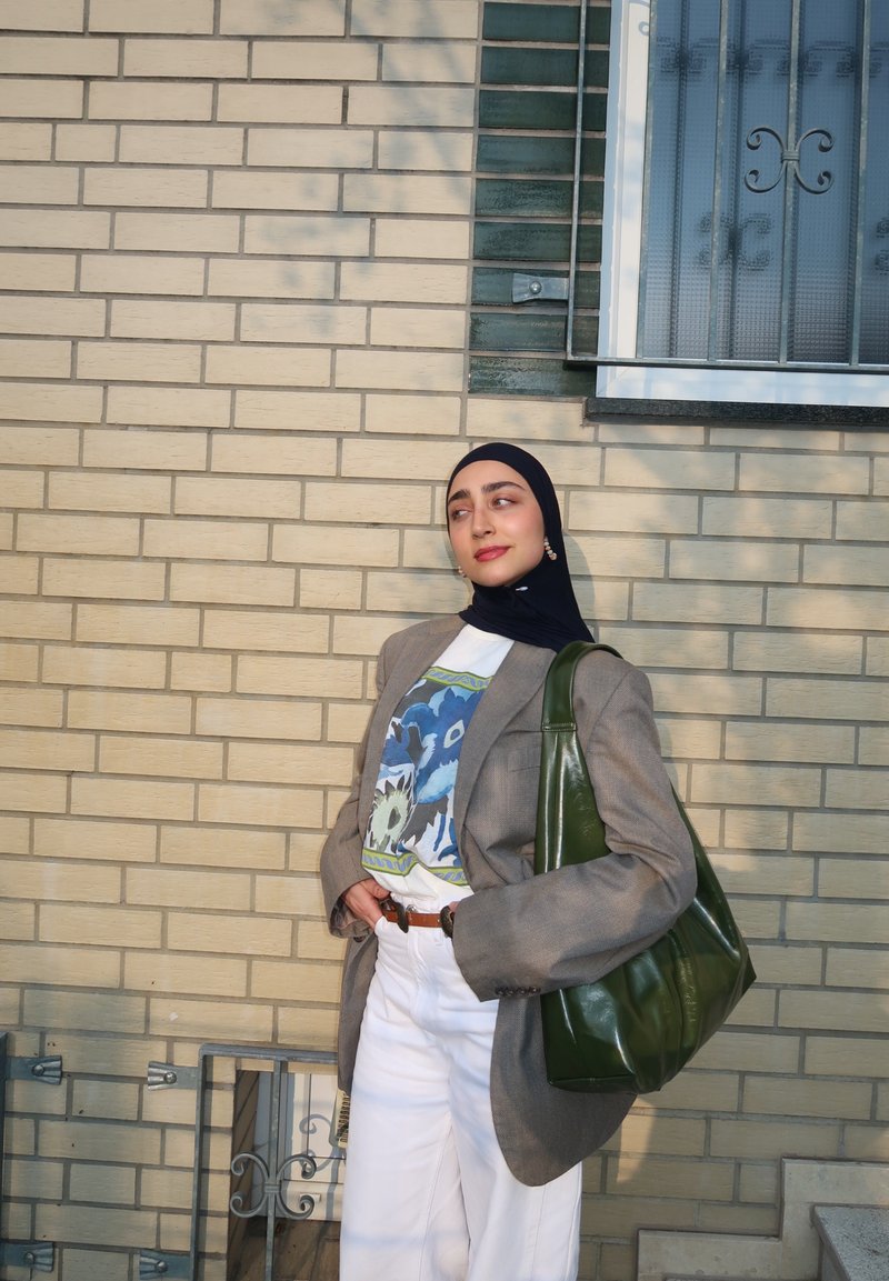 Jonge vrouw in een zwarte hijab draagt een grijze blazer, een grafisch T-shirt, witte broek en draagt een grote groene schoudertas tegen een bakstenen muur.