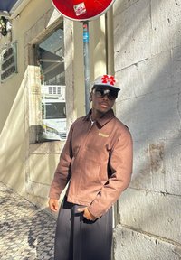 Veste marron à fermeture éclair avec col et poches lat�érales, portée avec un pantalon noir. Accessoires comprenant une casquette blanche avec un logo rouge et des lunettes de soleil.