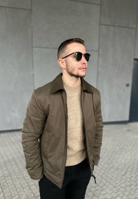 Ung mann iført solbriller, brun jakke, beige genser og svarte bukser som står på flislagt gulv med grå veggbehandling i bakgrunnen.