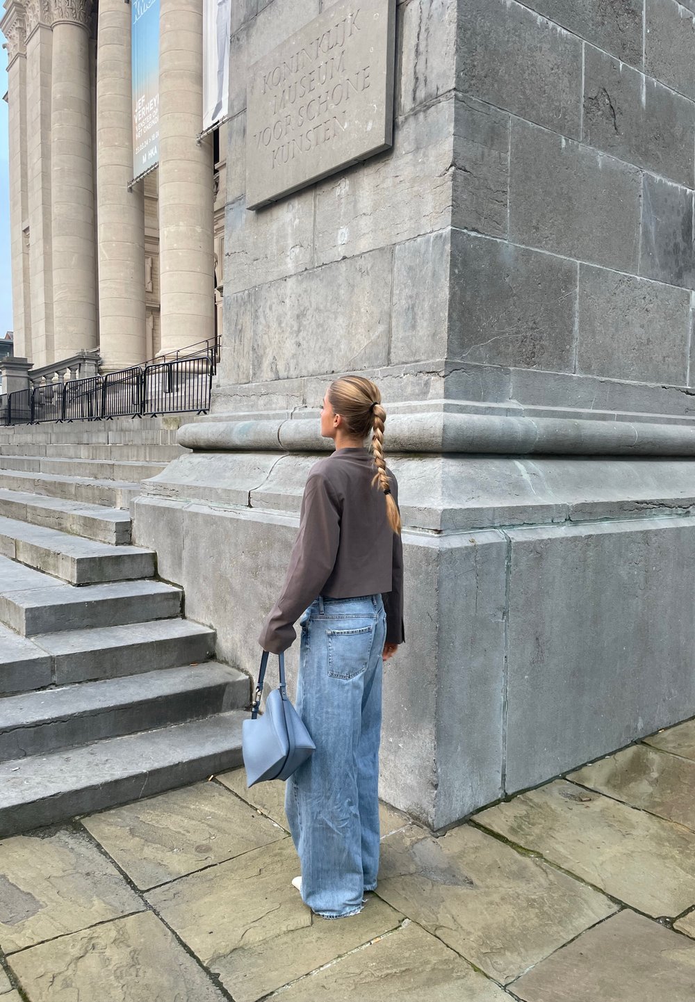 Wijdvallende denim jeans, een verkorte bruine jas en een blauwe handtas; stenen trappen en de gevel van het museum bieden een gestructureerde grijze achtergrond.
