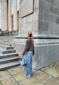 Wijdvallende denim jeans, een verkorte bruine jas en een blauwe handtas; stenen trappen en de gevel van het museum bieden een gestructureerde grijze achtergrond.
