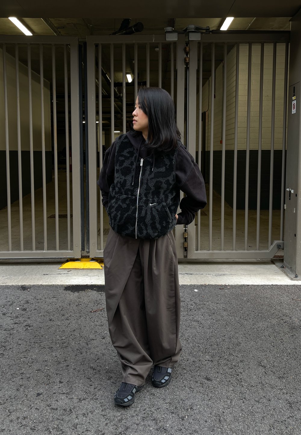 Sort fleecevest med mønster, båret over en mørk hættetrøje. Brune bukser med vide ben og sort fodtøj med detaljer. Urban baggrund med porte.