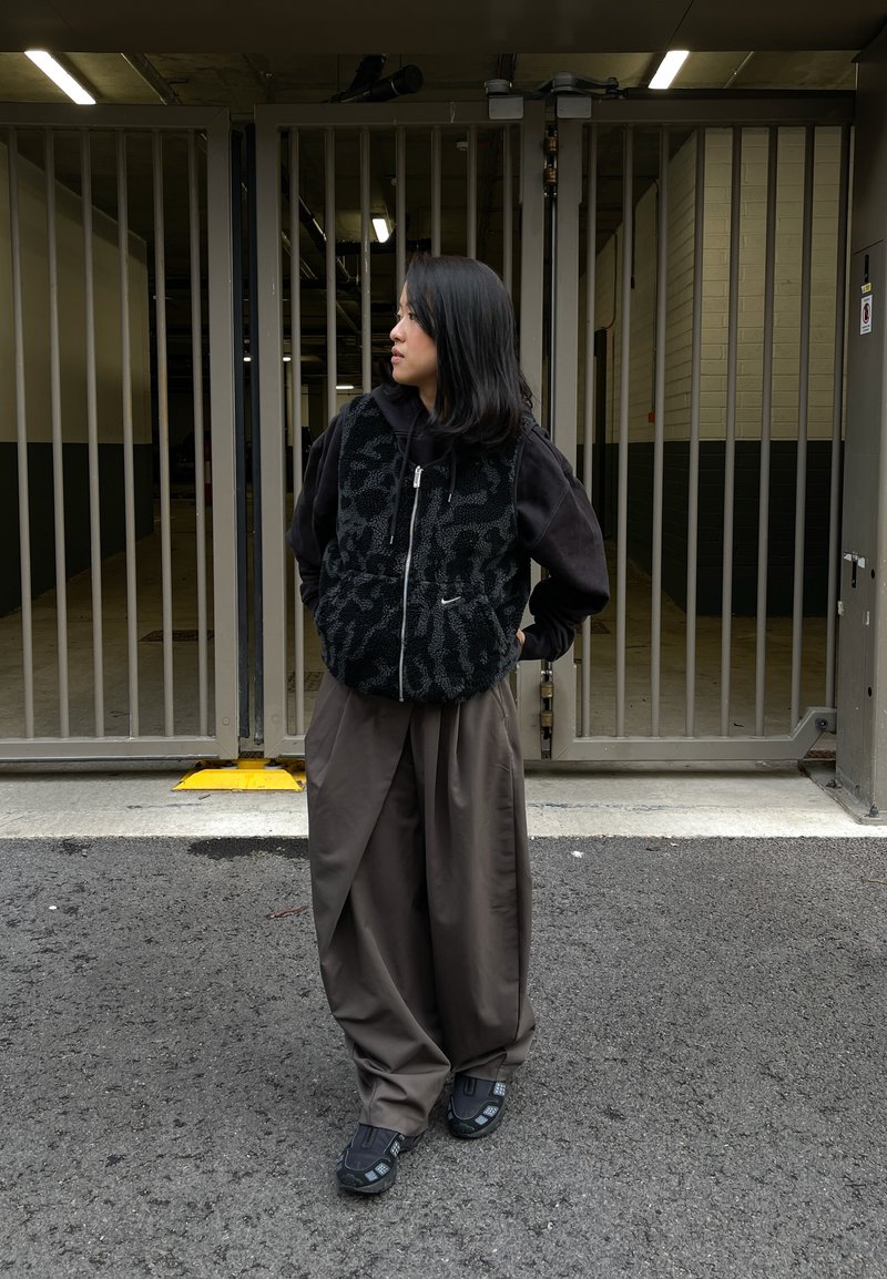 Gilet en polaire noire avec motif, porté par-dessus un sweat à capuche foncé. Pantalon marron à jambes larges et chaussures noires avec accents. Arrière-plan urbain avec des portails.