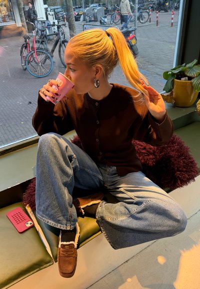 Cardigan marrón abotonado, vaqueros oversized azul claro, zapatillas marrones forradas de borrego. Sosteniendo una taza rosa. Cojín borroso de color granate cerca.