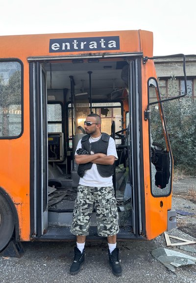 Autobús urbano naranja con las puertas abiertas, un hombre con pantalones cortos de camuflaje, chaleco negro, gafas de sol y botas negras está de pie en la puerta, en un entorno urbano.