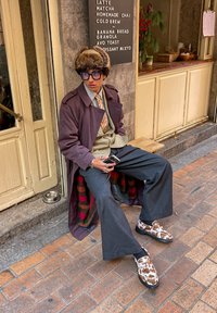 Υπερμεγέθη καφέ παλτό πάνω από γκρι ολόσωμο παντελόνι, με γκρι παντελόνι φαρδιάς γραμμής και παπούτσια καφέ-άσπρης ριγέ, καθισμένος σε μια καρώ κουβέρτα.