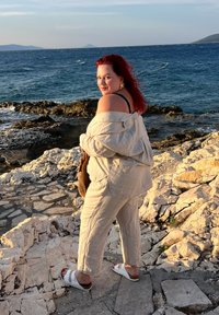 Lnianowy strój w jasnym beżu, bluzka z odkrytymi ramionami, pasujące spodnie, białe sandały. Stojąca na kamienistej linii brzegowej z niebieską wodą w tle.