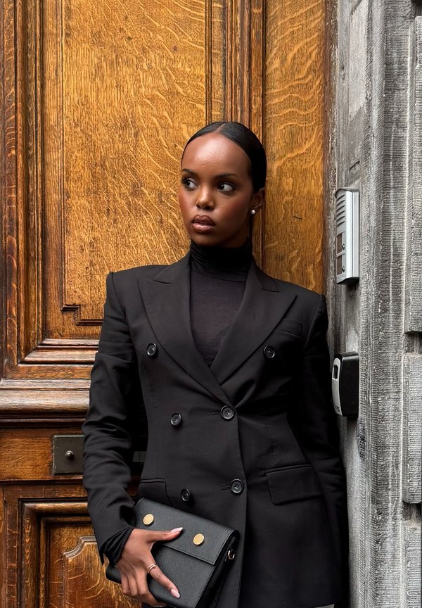 Schwarze doppelt geknöpfte Blazerjacke mit strukturierter Passform, getragen über einem Rollkragenpullover. Hält eine schwarze Clutch mit goldenen Akzenten. Holzhintergrund.