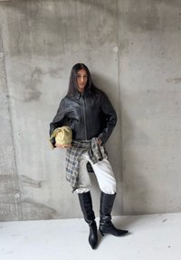 Veste en cuir noire avec col, pantalon blanc et bottes noires montant jusqu'aux genoux. Tient un sac beige. Fond en béton gris.