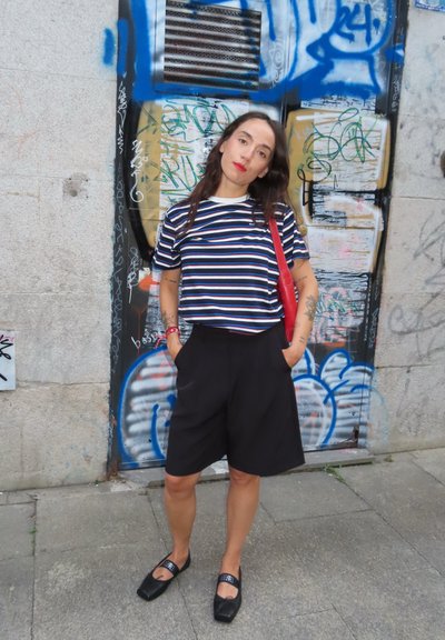 Camiseta de rayas blancas y negras con detalles rojos, pantalones cortos negros y zapatos negros. El fondo presenta graffiti en una pared.