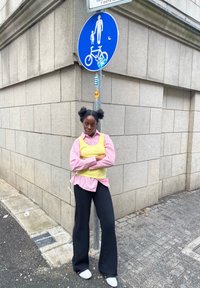 O persoană stă sprijinită de un zid de piatră, purtând o vestă galbenă peste o cămașă cu dungi roz și albe și pantaloni evazați negri, cu pantofi albi.