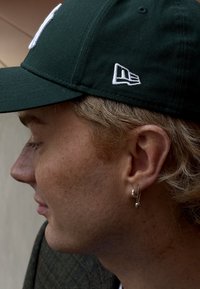Gorra de béisbol verde con un logo blanco en la parte frontal y un logo lateral. Pendiente de aro plateado visible en la oreja, piel con pecas, cabello texturizado.