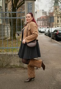 Femme en robe noire, veste marron clair et bottes en fourrure brune se tient sur un trottoir de la ville près d'une clôture en métal et de voitures garées.
