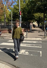 Olivově zelený svetr, šedé kalhoty širokého střihu, černá taška s třásněmi, kráčející po přechodu pro chodce. Viditelné stromy a Dopravní značky.