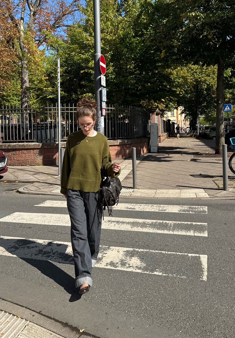 Pull vert olive, pantalon large gris, sac noir avec des franges, marchant sur un passage piéton. Des arbres environnants et des panneaux de signalisation sont visibles.