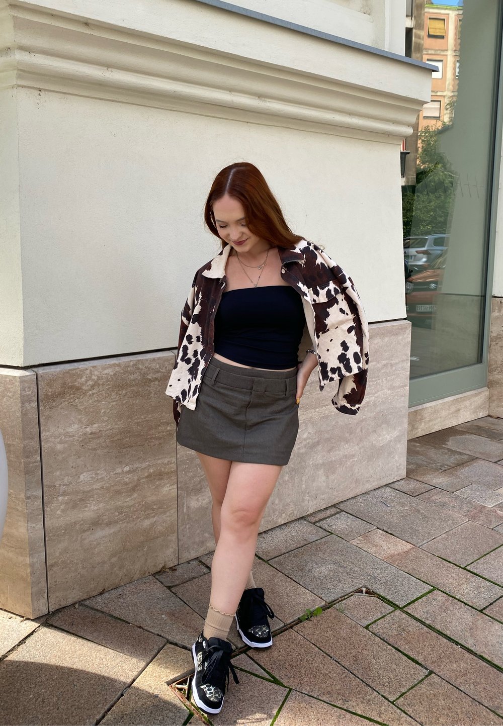 Brun og hvid mønstret jakke, sort strapless top, grå mini nederdel og sorte sneakers med hvide detaljer. Overfladerne er flisebelagte.