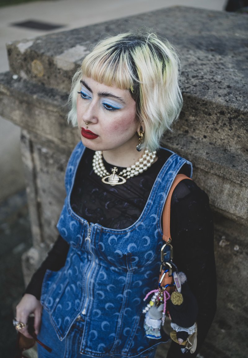 Denim vest with moon pattern, layered over a sheer black top. Short, wavy hair with pale highlights. Chunky pearl necklace and various keychains.