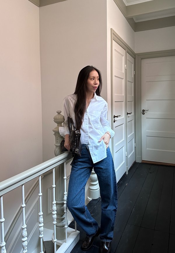 Una mujer con una camisa blanca y vaqueros azules se apoya en la barandilla de una escalera y lleva un bolso negro en un pasillo con puertas blancas.