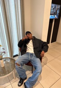 En mann iført svart jakke, beanie og revne jeans sitter avslappet i en stol ved et glassbord med en blomstervase i et moderne rom.