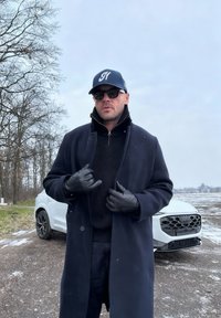 Άνδρας με σκούρο παλτό, δερμάτινα γάντια, γυαλιά ηλίου και καπέλο μπέιζμπολ στέκεται στο χιονισμένο δρόμο με ένα λευκό αυτοκίνητο και γυμνά δέντρα πίσω του.