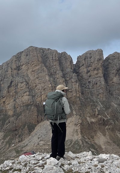 Mochila de material verde apagado, apoyada en terreno rocoso. Portador con chaqueta gris clara y gorra beige, mirando hacia un paisaje montañoso agreste.