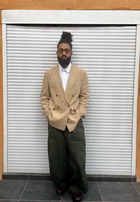 Sacou bej cu nasturi dubli purtat peste o cămașă albă, asortat cu pantaloni verzi cu croială largă și pantofi negri, pe fundal de jaluzea de argint.