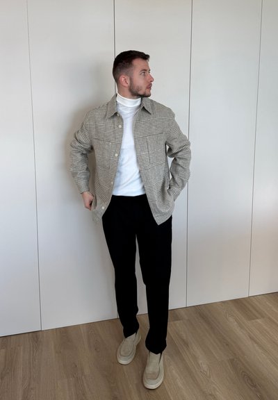 Hombre de pie en el interior, vestido con una chaqueta de cuadros, jersey de cuello alto blanco, pantalones negros y zapatos beige, mirando hacia su izquierda.