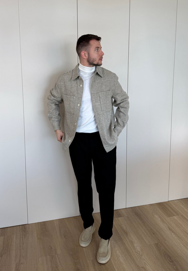 Άνδρας που στέκεται μέσα, φοράει καρό μπουφάν, λευκό ζιβάγκο, μαύρο παντελόνι και μπεζ παπούτσια, κοιτάει προς τα αριστερά.