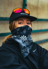 Bandana noire avec des motifs floraux et cachemire blancs, portée comme couverture du visage. Veste noire, lunettes de soleil sans monture avec des verres miroir, et casquette.