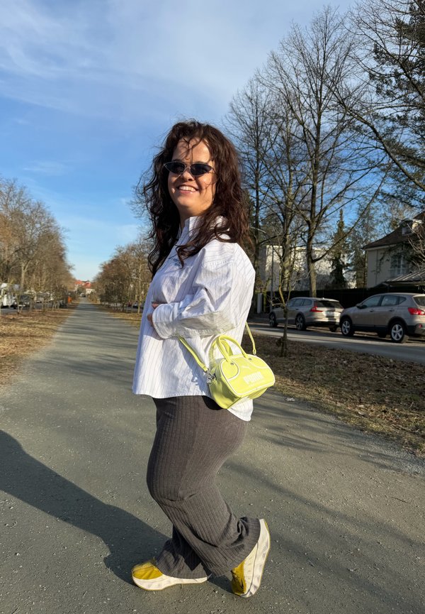 Sieviete ar saulesbrillēm, baltu kreklu, pelēkām biksēm, dzeltenām kurpēm, nēsā neona dzeltenu Puma somu, stāvot koku apzaļumotā ielā saulainā dienā.