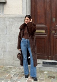 Long manteau en fausse fourrure marron avec de larges revers, associé à un col roulé marron, un jean évasé bleu et un petit sac à main marron. Bottines noires.