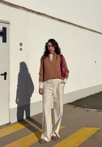 Kamizelka swetrowa w jasnobrązowym wełnianym materiale, biała koszula z kołnierzykiem pod spodem, kremowe szerokie spodnie oraz różowa torebka. Cień rzucony na chodnik.