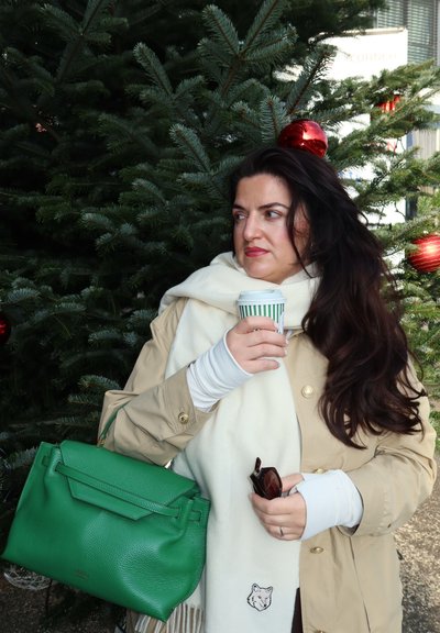 Abrigo trench beige, bufanda blanca con un detalle de zorro, bolso de cuero verde, sosteniendo una taza con franjas verdes, rodeado de ramas de pino y adornos rojos.