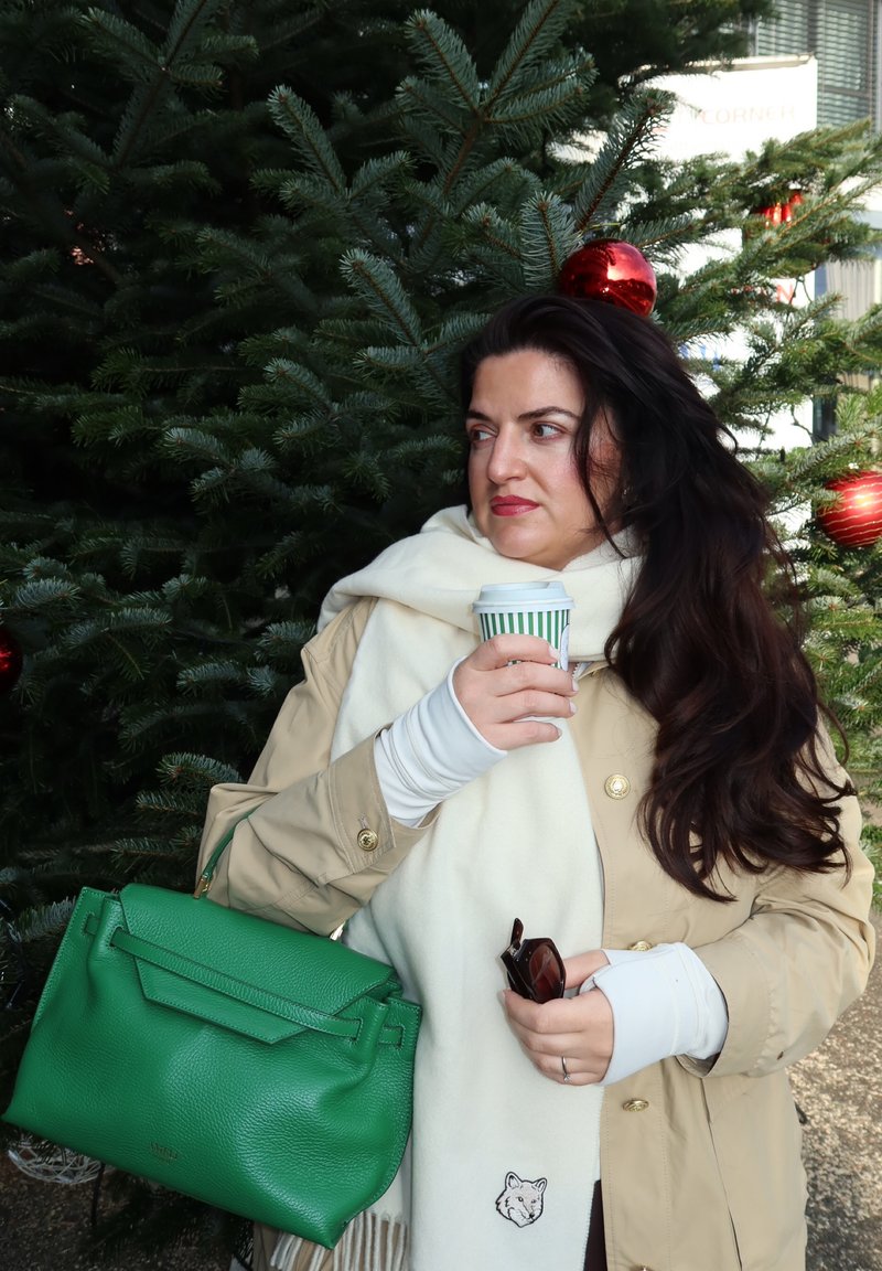 Beiger Trenchcoat, weißer Schal mit einem Fuchsdetail, grüne Ledertasche, hält eine Tasse mit grünen Streifen, umgeben von immergrünen Zweigen und roten Schmuckstücken.