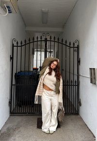 Henkilö, jolla on päällään kermaan vivahtava palmikkoneule, oliivinvihreä takki, valkoiset leveälahkeiset housut ja beige ruudullinen huivi. Seisoo mustan rauta-aidan vieressä.