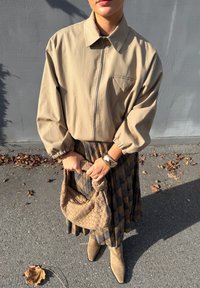 Beige ritssluitingjack met pofmouwen, gecombineerd met een geplooide rok met een geruit patroon. Houdt een gevlochten beige tas vast en draagt beige laarzen.