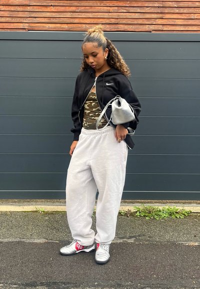 Chaqueta negra de Nike con cremallera sobre una camiseta de tirantes de camuflaje, combinada con pantalones de chándal grises y zapatillas plateadas con acentos rojos. Bolsa gris en la mano.
