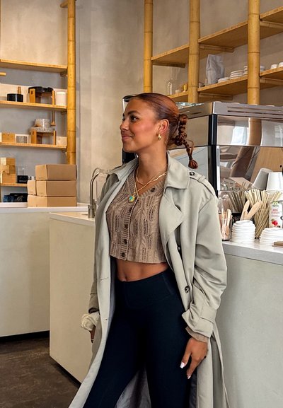 Un abrigo trench gris claro sobre una blusa cropped beige con un patrón texturizado, combinado con leggings negros de cintura alta, en el interior de un café moderno.