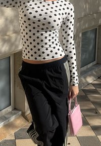 Personne portant un haut à manches longues blanc à pois et un pantalon de survêtement noir, tenant un petit sac à main rose sur un trottoir carrelé.