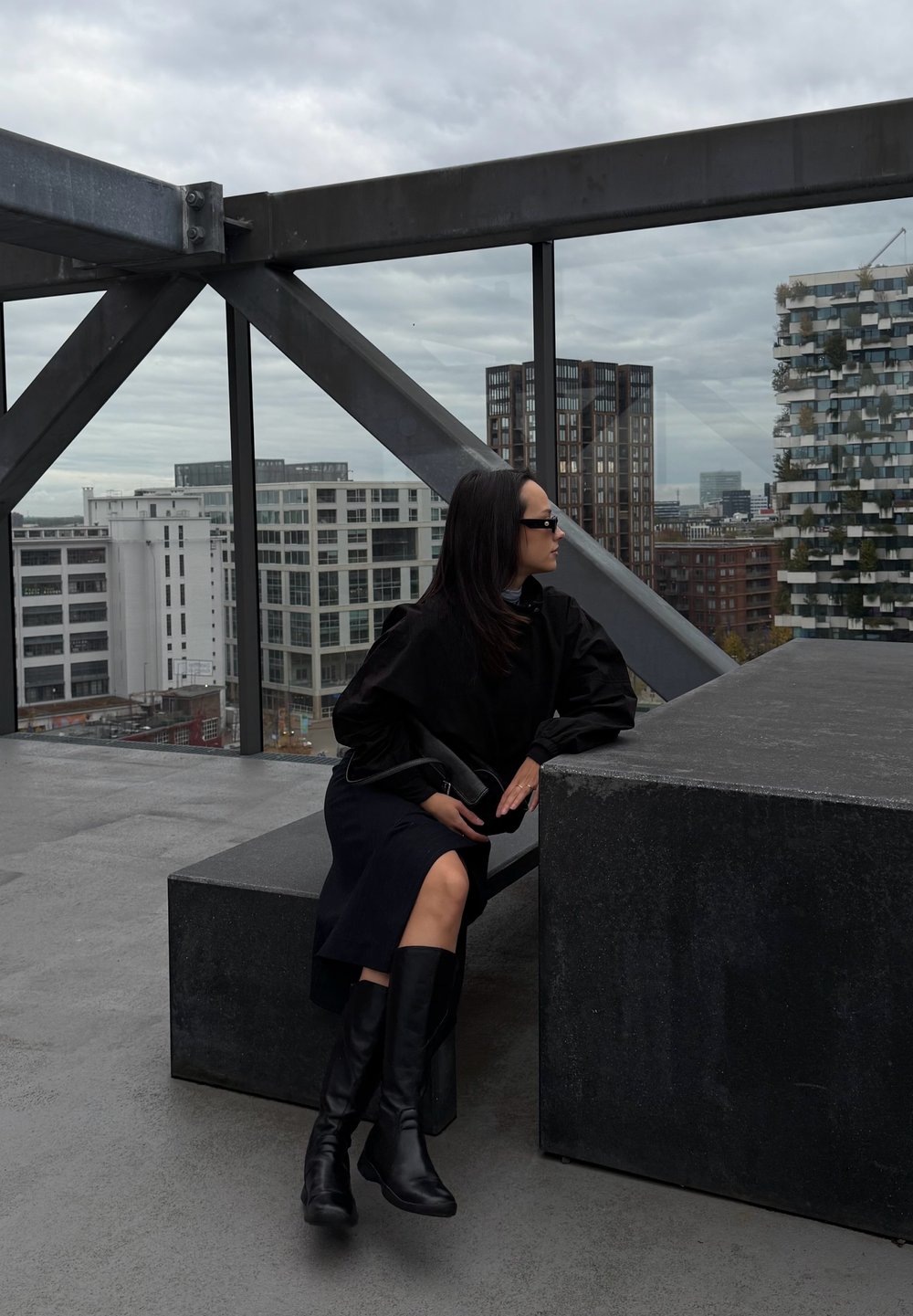Top oversized preto com mangas volumosas, saia preta e botas pretas até ao joelho. Assentos cinzentos num terraço com fundo urbano e céu nublado.