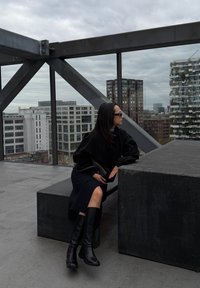 Top oversized preto com mangas volumosas, saia preta e botas pretas até ao joelho. Assentos cinzentos num terraço com fundo urbano e céu nublado.