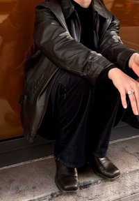 Melnā ādas jaka virs melnas topiņas, kopā ar tumšām džinsa biksēm un brūniem potīšu zābakiem. Vienā rokā sudraba gredzens ar ziedu detaļu.