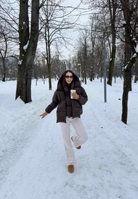 Femme en doudoune sombre, pantalon beige et gants, tenant une tasse, debout sur une jambe sur un sentier enneigé du parc bordé d'arbres nus.