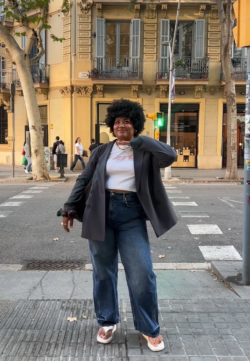 Blazer nero oversize, t-shirt bianca corta con logo, jeans blu a vita alta e infradito bianche, in piedi su una strada della città.