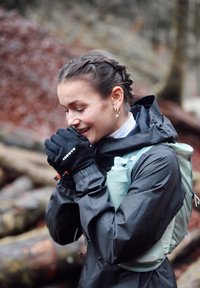 Gants noirs avec logo, veste imperméable et sac d'hydratation vert clair. Cheveux tressés et décor naturel avec feuillage flou.