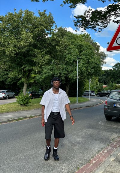 Persona con una camiseta blanca de manga corta, pantalones cortos negros y botas negras, caminando por una carretera bordeada de árboles y coches aparcados.