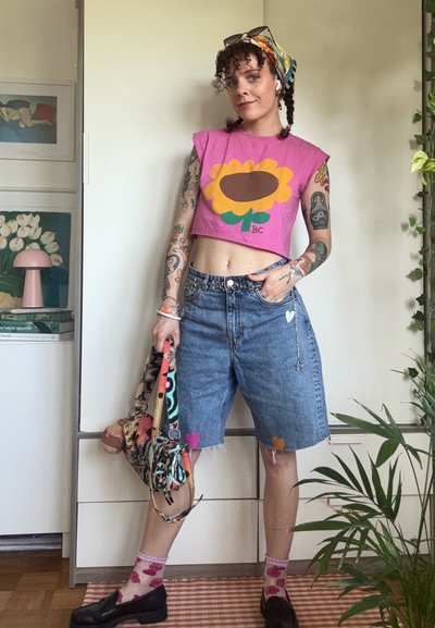 Joven mujer con cabello rizado y tatuajes lleva un top corto rosa, shorts vaqueros con parches de corazones, calcetines estampados, zapatos negros y sostiene una bolsa colorida.