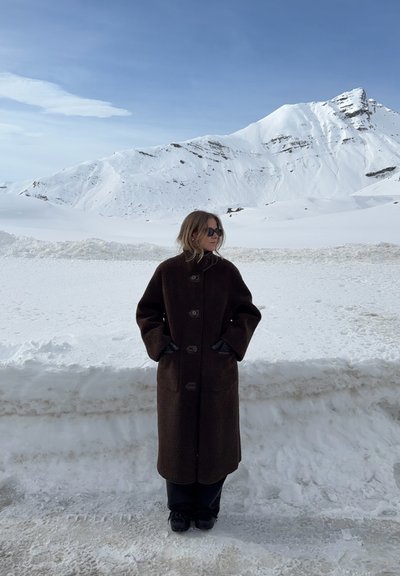 Persona con abrigo oscuro y guantes de pie sobre suelo nevado con montañas cubiertas de nieve y cielo azul de fondo.