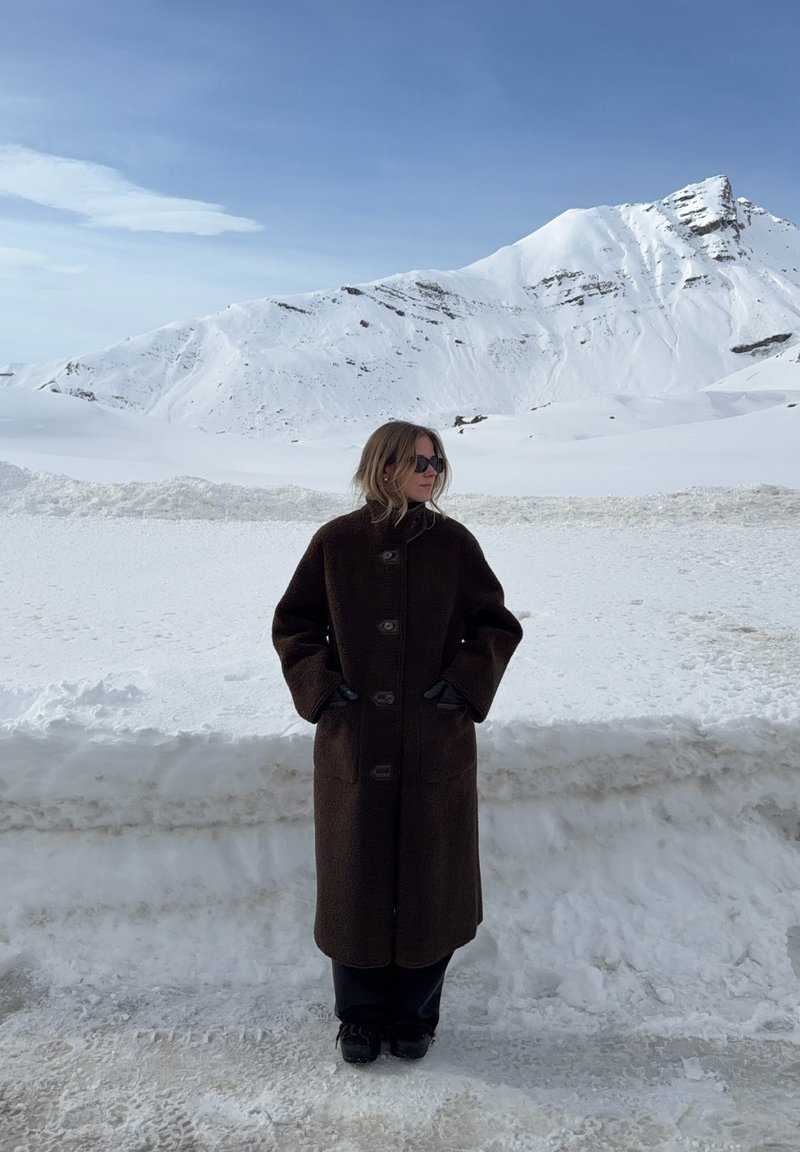 Person i mørk frakke og handsker står på snedækket jord med sneklædte bjerge under blå himmel i baggrunden.